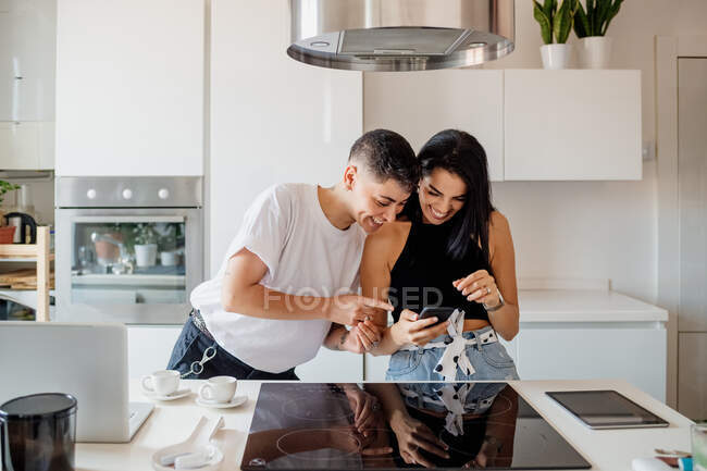 Joven pareja lesbiana de pie en la cocina, mirando el teléfono móvil - foto de stock