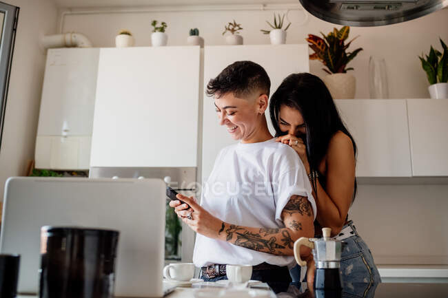 Young lesbian couple standing in kitchen, looking at mobile phone — Stock Photo