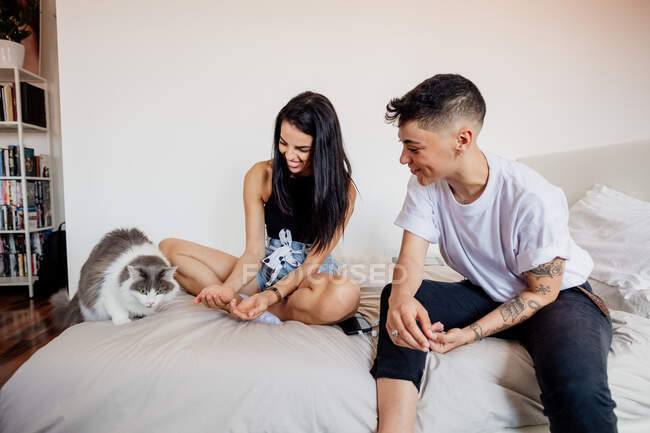 Jovem casal lésbico sentado em uma cama, brincando com o gato. — Fotografia de Stock