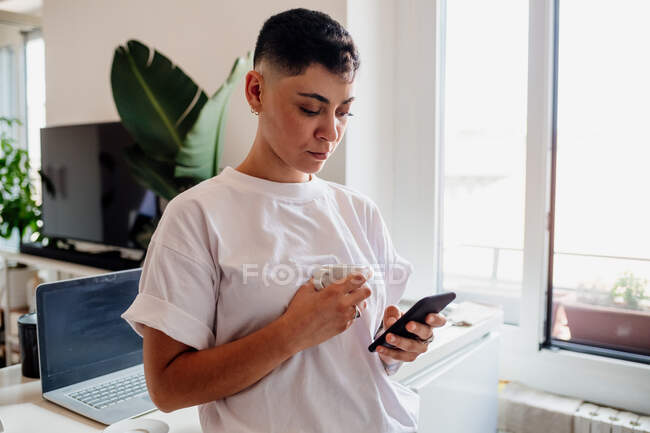 Young woman with shaved head standing in a kitchen, checking her mobile phone — Stock Photo