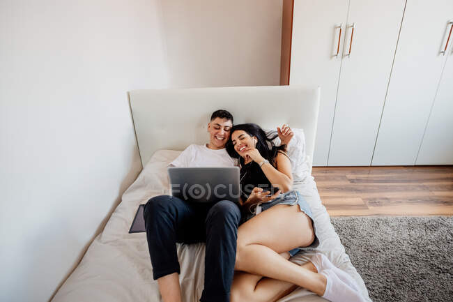 Junges lesbisches Paar liegt auf einem Bett und schaut auf Handy und Laptop — Stockfoto