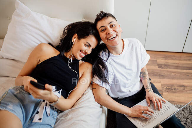 Молодая лесбийская пара в спальне, держит мобильный телефон и ноутбук, улыбается в камеру — стоковое фото