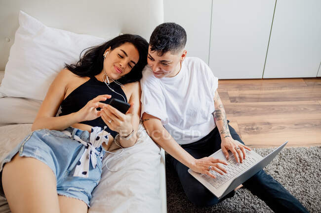 Giovane coppia lesbica in una camera da letto, utilizzando il telefono cellulare e laptop — Foto stock