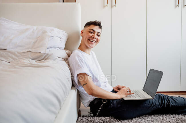 Молода жінка з поголеною головою сидить у спальні, використовуючи ноутбук, посміхаючись на камеру — стокове фото