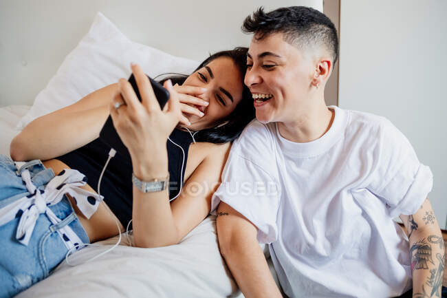 Pareja lesbiana joven en un dormitorio, mirando el teléfono móvil, sonriendo - foto de stock