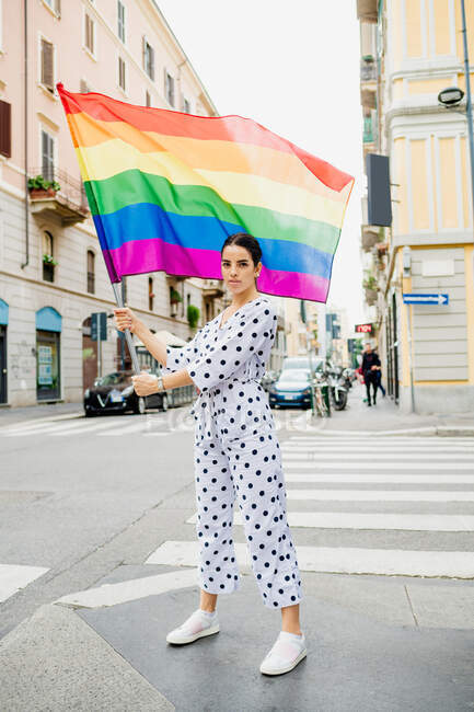 Mujer lesbiana joven de pie en una calle, ondeando la bandera del arco iris. - foto de stock