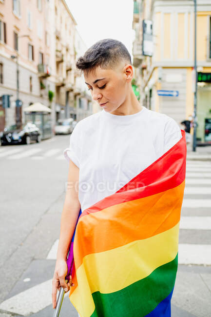 Jovem lésbica de pé em uma rua, envolto em bandeira do arco-íris — Fotografia de Stock