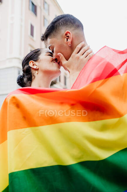 Portrait of young lesbian couple standing on a street, wrapped in rainbow flag, kissing — Stock Photo