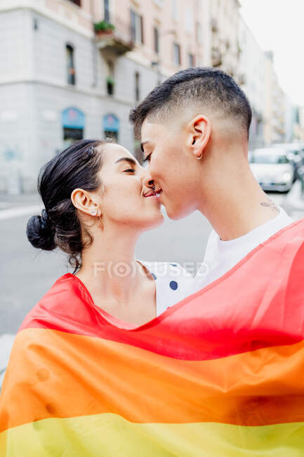 Portrait of young lesbian couple standing on a street, wrapped in rainbow flag, kissing — Stock Photo