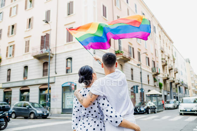 Rear view of young lesbian couple standing on a street, waving rainbow flag — Stock Photo