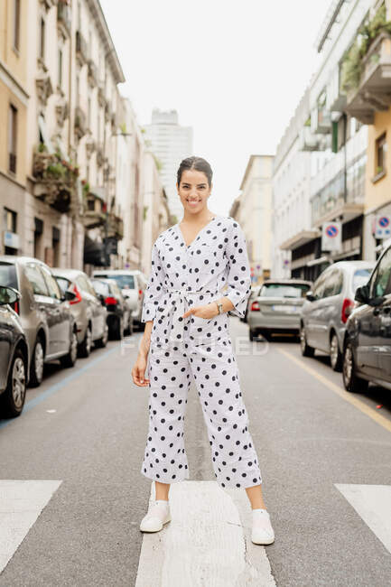Portrait of smiling woman wearing white and black polka dot jumpsuit, standing on pedestrian crossing — Stock Photo