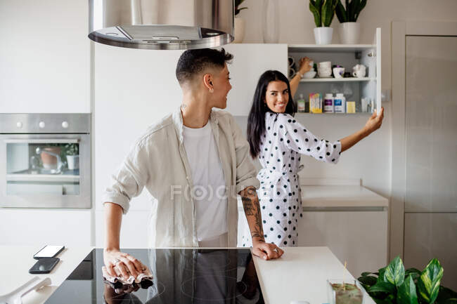 Joven pareja lesbiana de pie en la cocina, sonriéndose mutuamente. - foto de stock