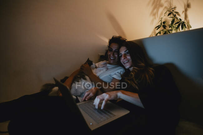 Las mujeres lesbianas relajarse en el sofá y el uso de la computadora portátil por la noche - foto de stock