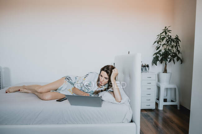 Жінка з коричневим волоссям лежить на білому ліжку, дивлячись на ноутбук . — стокове фото