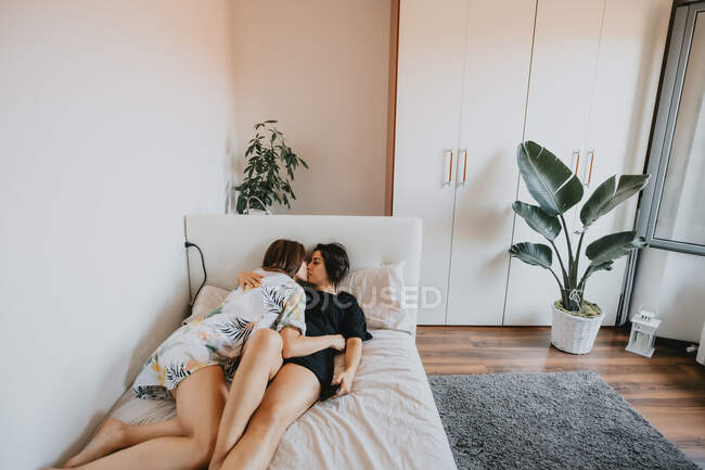 Two women with brown hair lying on white daybed, hugging and kissing. — Stock Photo