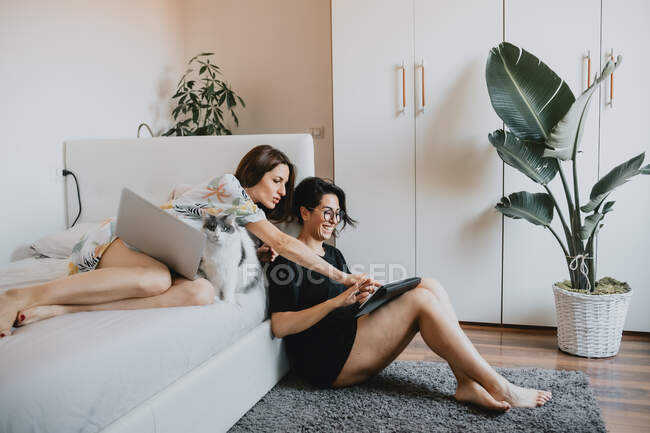 Две женщины с коричневыми волосами сидят на полу и лежат на кровати, используя ноутбук и цифровой планшет. — стоковое фото