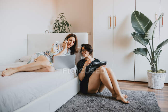 Two women with brown hair sitting on floor and lying on daybed, using laptop and mobile phone. — Stock Photo