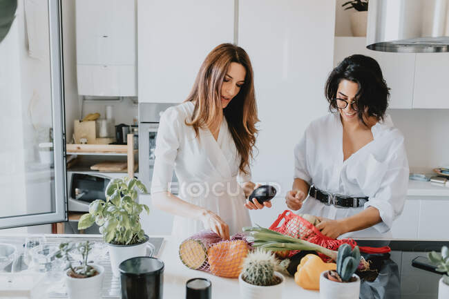 Две улыбающиеся женщины с каштановыми волосами стоят на кухне, удаляя овощи из сети магазинов. — стоковое фото