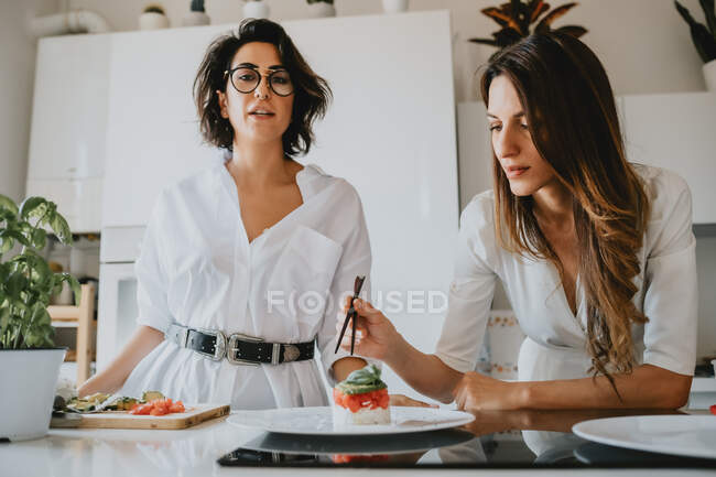 Dos mujeres sonrientes con el pelo castaño de pie en una cocina, preparando comida. - foto de stock
