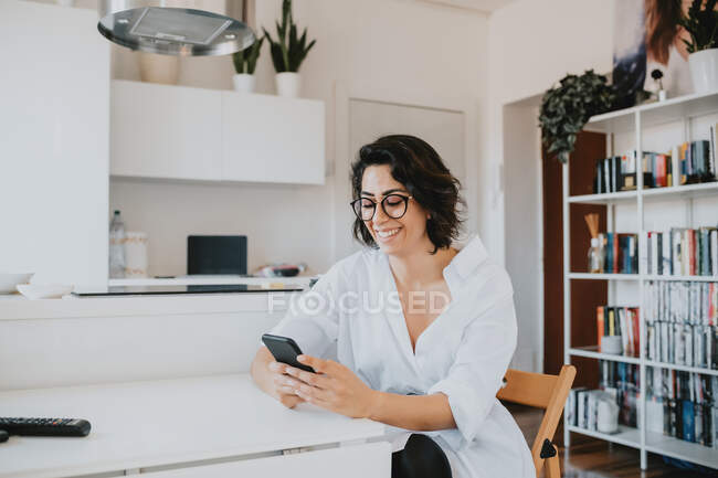 Frau mit braunen Haaren und Brille sitzt in einer Wohnung am Tisch und benutzt Handy. — Stockfoto