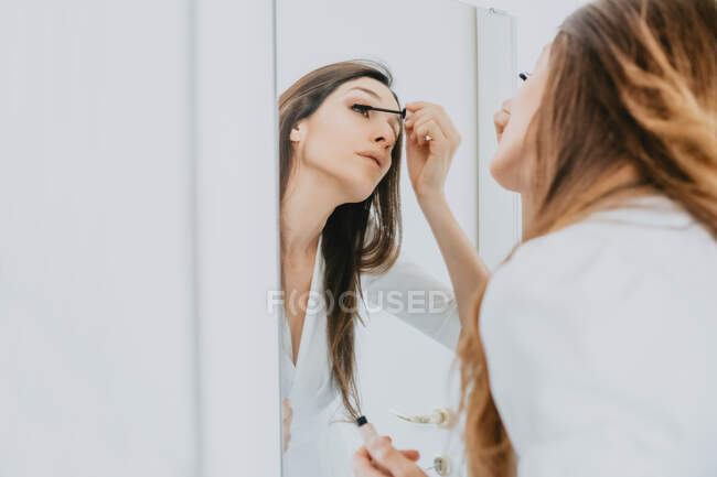 Frau mit braunen Haaren steht vor Spiegel und trägt Mascara auf. — Stockfoto