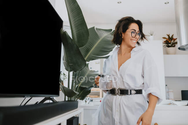 Mulher sorridente com cabelo castanho vestindo óculos de pé em um apartamento, regando planta. — Fotografia de Stock