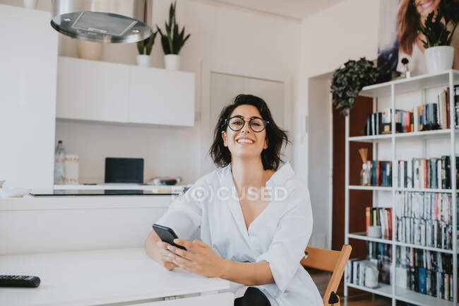 Frau mit braunen Haaren und Brille sitzt in einer Wohnung am Tisch und lächelt in die Kamera. — Stockfoto