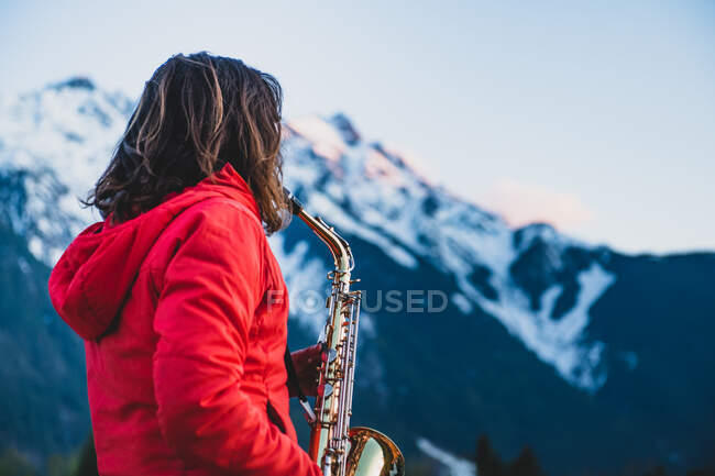 Женщина в ярко-красной куртке, играющая на саксофоне, заснеженная гора на заднем плане, Британская Колумбия, Канада — стоковое фото