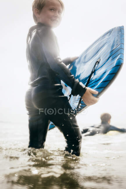 Porträt eines kleinen Jungen im Neoprenanzug, der Surfbretter ins Meer trägt, Santa Barbara, Kalifornien, USA. — Stockfoto
