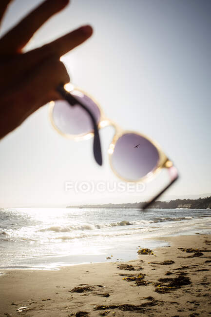 Primo piano di persona sulla spiaggia di sabbia, tenendo gli occhiali da sole verso il sole. — Foto stock
