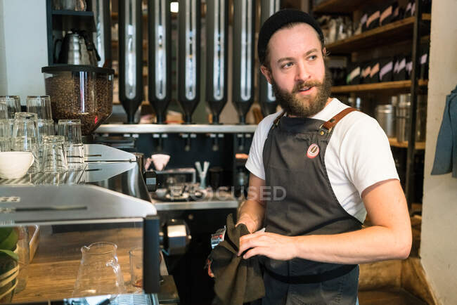 Bärtiger Barista mit Schürze arbeitet hinter dem Tresen in einem Café. — Stockfoto
