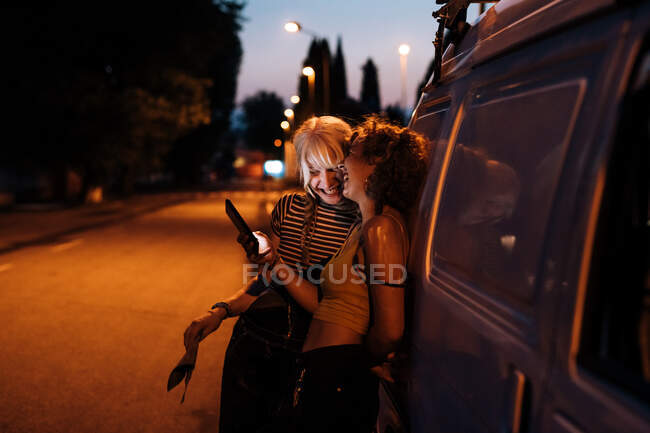 Pareja femenina riéndose con el teléfono por la noche, apoyada en la furgoneta - foto de stock