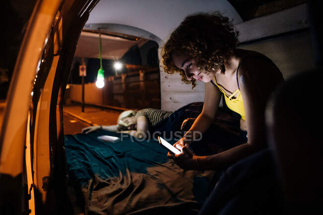 Jovem mulher olhando para o telefone como parceiro dorme na parte de trás da van — Fotografia de Stock