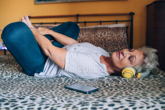 Старша жінка розслабляється на ліжку, слухаючи навушники — стокове фото