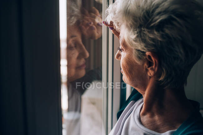 Mulher sênior olhando através da janela do apartamento — Fotografia de Stock