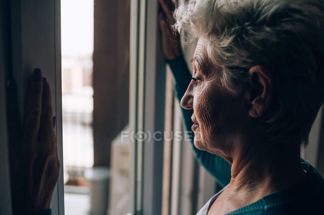 Mulher sênior olhando para fora da janela do apartamento — Fotografia de Stock