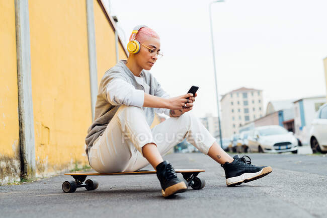 Young woman sitting on skateboard, wearing headphones, using cellphone — Stock Photo