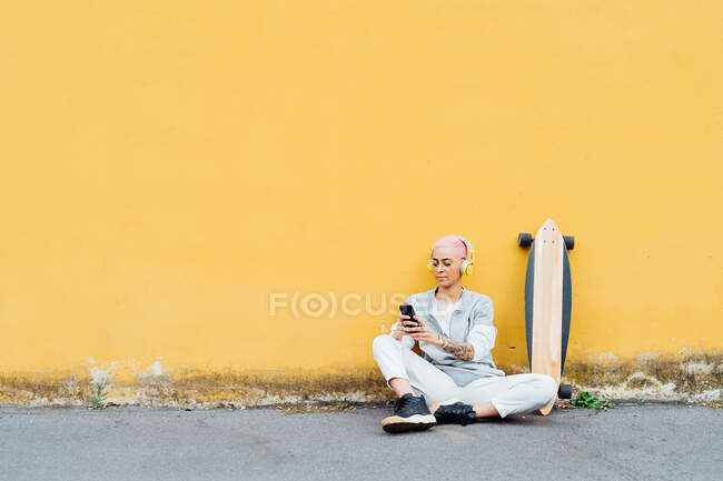 Скейтбордист сидить на підлозі перед жовтою стіною, використовуючи мобільний телефон — стокове фото
