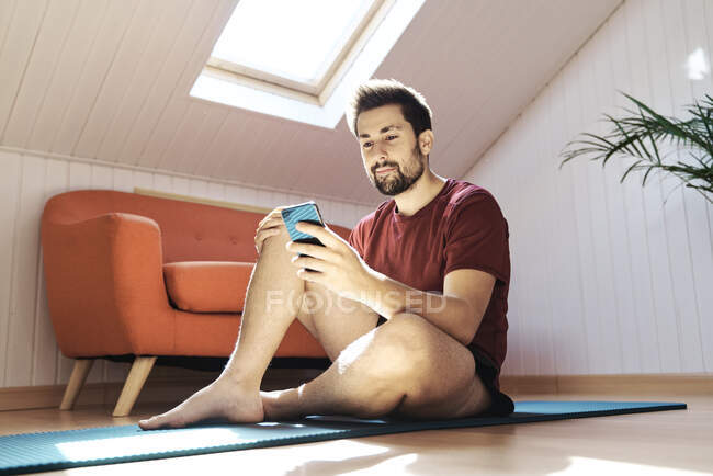 Чоловік сидить на тренувальному килимку, дивлячись на телефон — стокове фото
