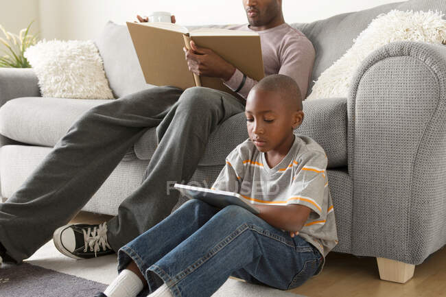 Хлопчик сидить на підлозі за допомогою цифрового планшета, батько читає книгу — стокове фото