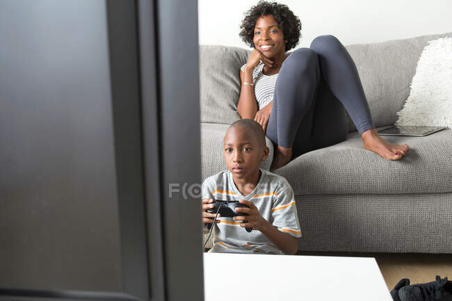 Junge spielt Videospiel, Mutter schaut vom Sofa aus zu — Stockfoto
