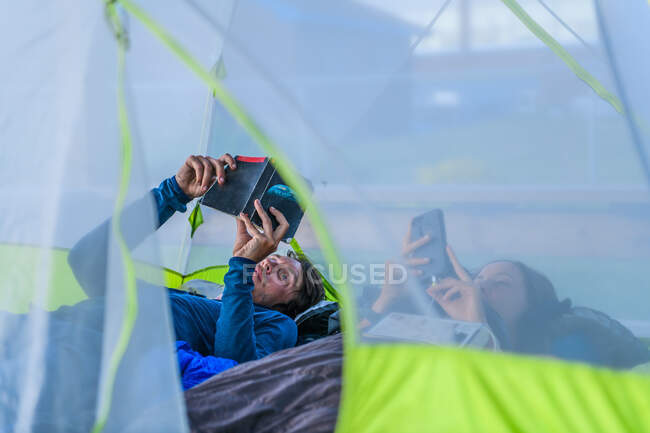 Paar im Zelt, liest und schaut aufs Handy — Stockfoto