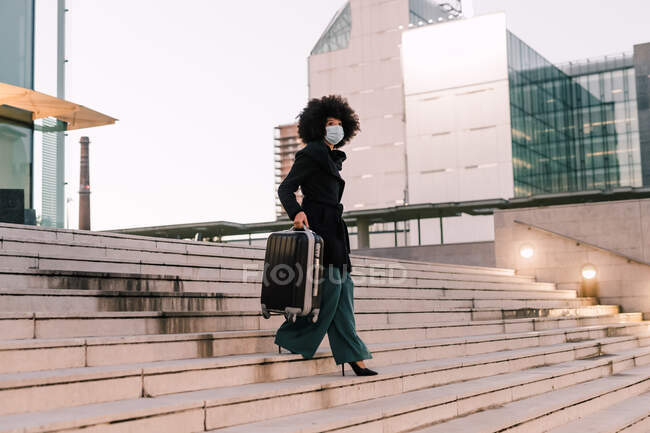 Businesswoman descending stairs with suitcase, wearing face mask — Stock Photo