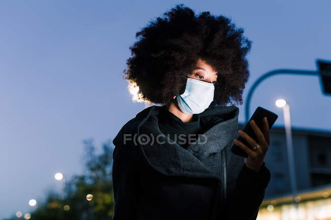 Woman wearing face mask and looking at phone, outdoors at night — Stock Photo