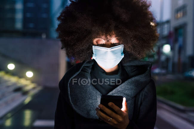 Woman wearing face mask, looking at mobile phone, illuminated at — Stock Photo