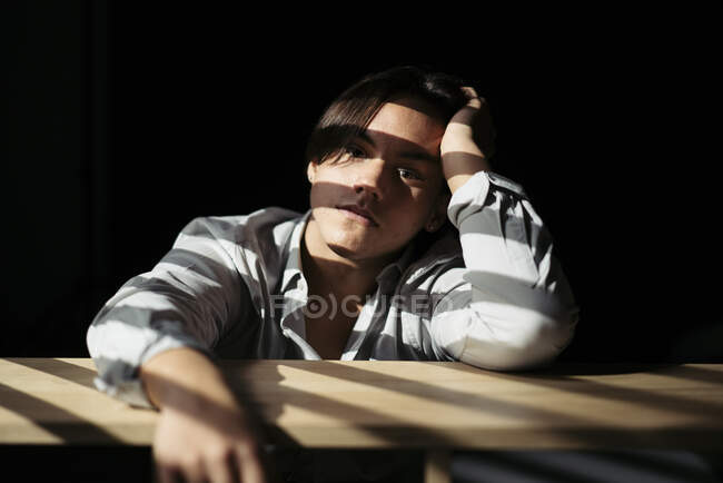 Teenagerjunge sitzt am Tisch in dunklem Interieur mit Sonnenlicht — Stockfoto