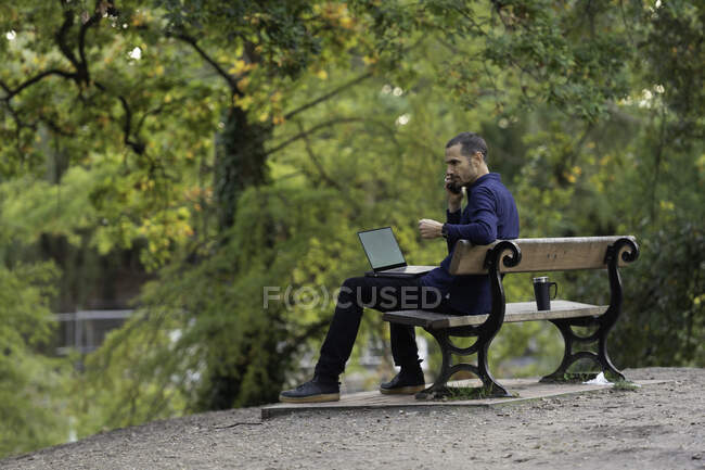 Homme sur banc de parc, prendre appel et travailler sur ordinateur portable — Photo de stock
