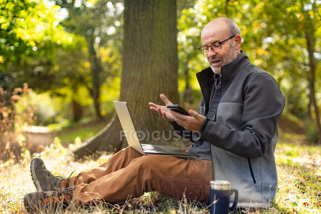 Человек сидит в парке и работает с ноутбуком и телефоном — стоковое фото