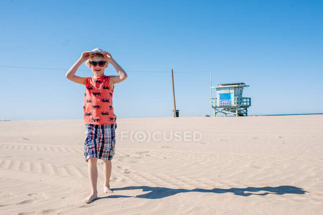 Boy on sunny sandy beach, wearing sunglasses and hat — Stock Photo