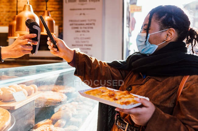 Italia, Mujer con mascarilla de pie pagando con un teléfono inteligente para merendar en la calle - foto de stock